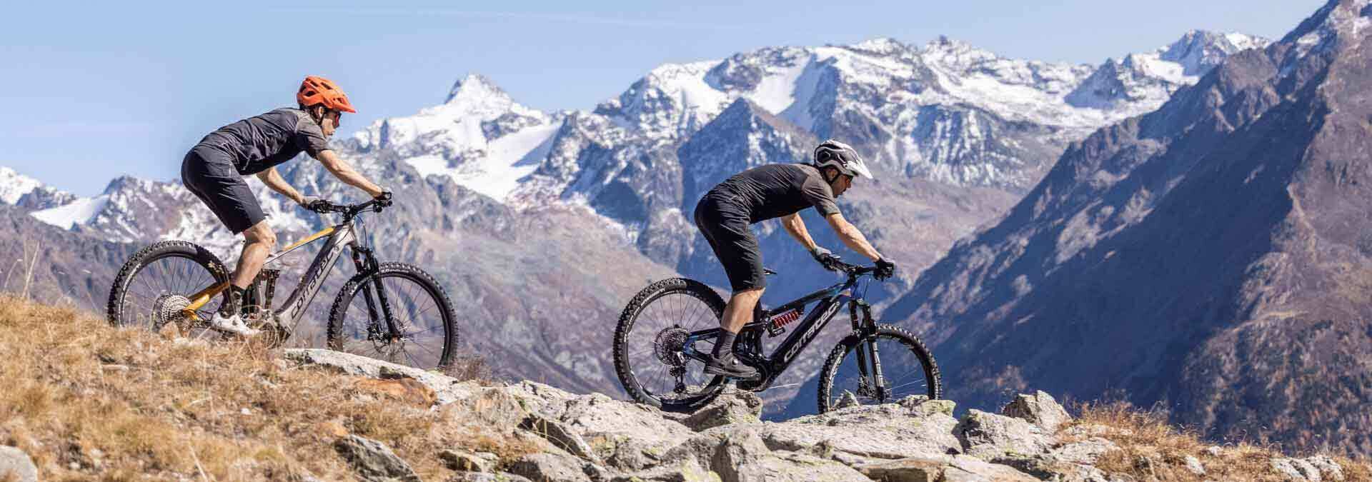 frau-und-mann-in-den-Bergem-fahren-auf-e-mountainbikes-fahren-bergab frau-und-mann-in-den-Bergem-fahren-auf-e-mountainbikes-fahren-bergab